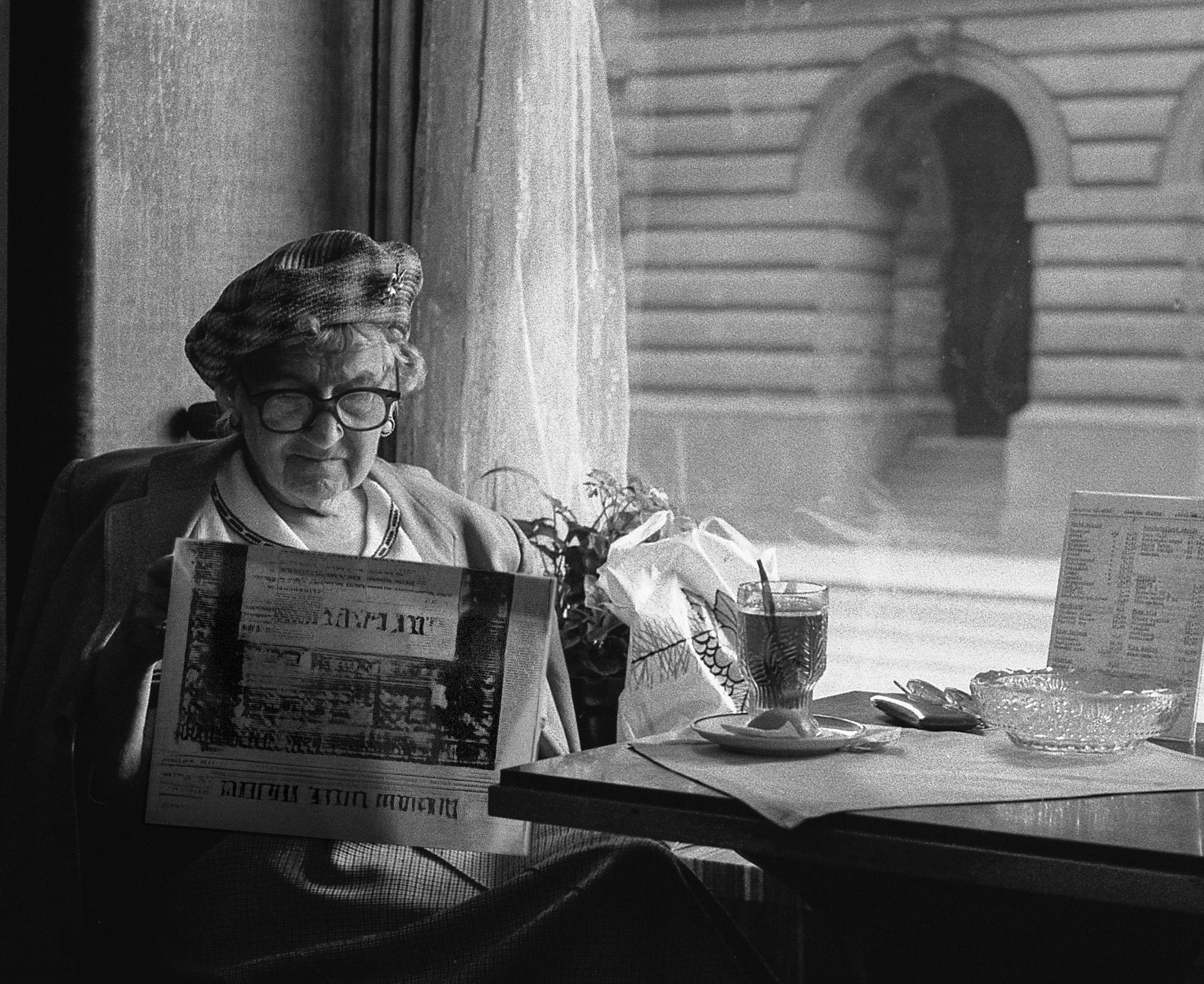old woman in glasses and a beret sitting at a table, reading a fake newspaper